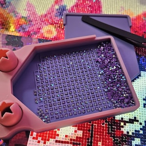 Puede incluir: Una bandeja de pintura de diamante de color morado claro con dos recortes en forma de estrella a la izquierda, llena de pequeños brillantes cuadrados morados dispuestos en cuadrícula. Unas pinzas de metal negro descansan sobre una tapa a juego.