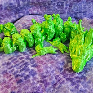 Puede incluir: Una figura de dragón articulada de color verde lima brillante que presenta una cabeza detallada con crestas prominentes y un cuerpo segmentado y flexible con escamas texturizadas. La criatura está en una posición curva, mostrando su intrincado diseño impreso en 3D.