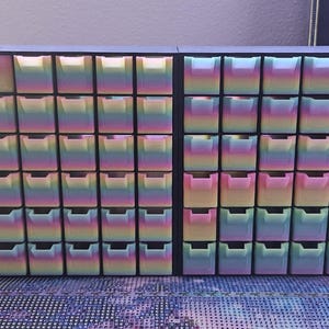 以下が含まれることがあります： 虹色のグラデーション仕上げが施された小さな引き出しのグリッドを備えた黒い収納オーガナイザー。このユニットは、複数の行と列の引き出しを持つ2つのセクションが並んで構成されており、側面にはいくつかのツールを収納するコンパートメントがあります。