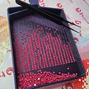 May include: A dark blue diamond painting tray filled with rows of small, square, red gems. Black tweezers rest on the tray. The tray has a handle and is partially filled with loose gems. The background has the word "huacan".