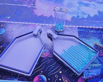 May include: Two white plastic diamond painting trays with blue edges, one empty and one containing rows of small light blue square resin drills. A clear plastic storage vial with a lid labeled 55 P 3846 sits between them, filled with light blue drills.