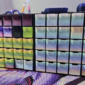 May include: A black storage unit with numerous small, coloured compartments. The compartments are arranged in rows and columns, with a variety of pastel shades. The unit is designed for small craft supplies, such as beads or diamond painting gems.