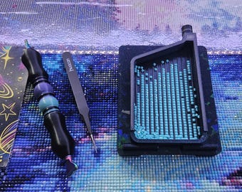 Op de afbeelding: Een diamond painting gereedschapsset bestaande uit een zwart-blauwe kralenpen, een zwarte pincet met de merknaam Huacan aan de zijkant, en een zwart gegroefd bakje met talloze kleine, vierkante, lichtblauwe harssteentjes.