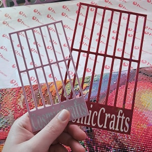 May include: Two plastic diamond painting tools. One is light pink, the other is red. Both have a grid pattern and the text "Cosmic Crafts" in white. The tools are held over a colorful diamond painting.