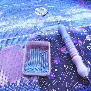 May include: A diamond painting kit featuring a light purple ergonomic stylus, a small rectangular tray filled with light blue square resin drills, and a clear plastic storage container, all set against a dark fabric with a space-themed print.