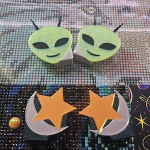 Puede incluir: Dos cabezas de alienígena de color verde claro con ojos negros y antenas, junto con dos formas de estrella y luna creciente de color naranja. Estos objetos tienen una superficie texturizada y están dispuestos en dos conjuntos sobre un fondo de rejilla.