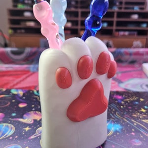 May include: White paw-shaped ceramic holder with three colorful glass tools. The tools have iridescent finishes in pink, blue, and light blue.
