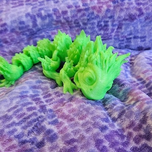 Puede incluir: Una figura de lagarto articulada de color verde lima brillante con una superficie texturizada y puntiaguda. La criatura presenta un diseño de cuerpo segmentado que permite el movimiento, con una cabeza detallada, ojos visibles y pies con garras.