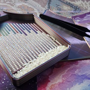 Pode incluir: Uma bandeja multicamadas para pintura de diamantes com um bico em forma de funil, contendo inúmeras pequenas contas quadradas brancas dispostas em canais paralelos. Uma pinça de metal preto repousa sobre uma ferramenta plana, escura e iridescente.