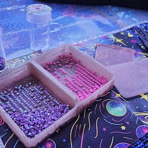 Pode incluir: Close-up de materiais de pintura de diamantes. Dois pequenos frascos de plástico transparente cheios de pedras roxas estão ao lado de uma bandeja dividida contendo pedras rosas e roxas. A bandeja e os frascos estão em uma superfície estampada.