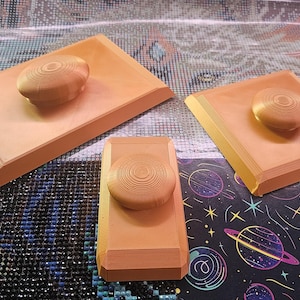 Può includere: Tre fermi per diamond painting di colore arancione chiaro, di forma rettangolare e quadrata, con una manopola circolare centrale. Ogni strumento presenta una base piatta e un'impugnatura testurizzata a coste per fissare la pellicola protettiva.