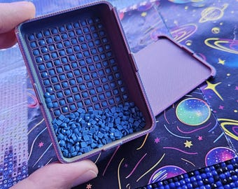 May include: A rectangular purple plastic tray with grooved channels containing numerous small, square, dark blue diamond painting drills. The tray has a matching detachable lid resting nearby, both featuring a textured, cross-hatched surface finish.