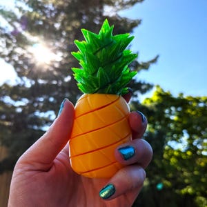 May include: A 3D printed pineapple toy with a yellow body and green leaves. The pineapple is being held in a hand.