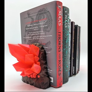 May include: A bookend featuring a red crystal rock formation and a dark brown wooden base. The bookend is holding up a stack of books with the titles "A Court of Thorns and Roses" and "A Court of Mist and Fury" by Sarah J. Maas.