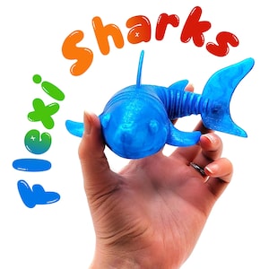 Może przedstawiać: Niebieska zabawka w kształcie rekina z płetwami i ogonem trzymana w dłoni. Słowo "sharks" jest napisane na pomarańczowo i czerwono nad rekinem. Słowo "flexi" jest napisane na zielono i niebiesko po lewej stronie obrazu.