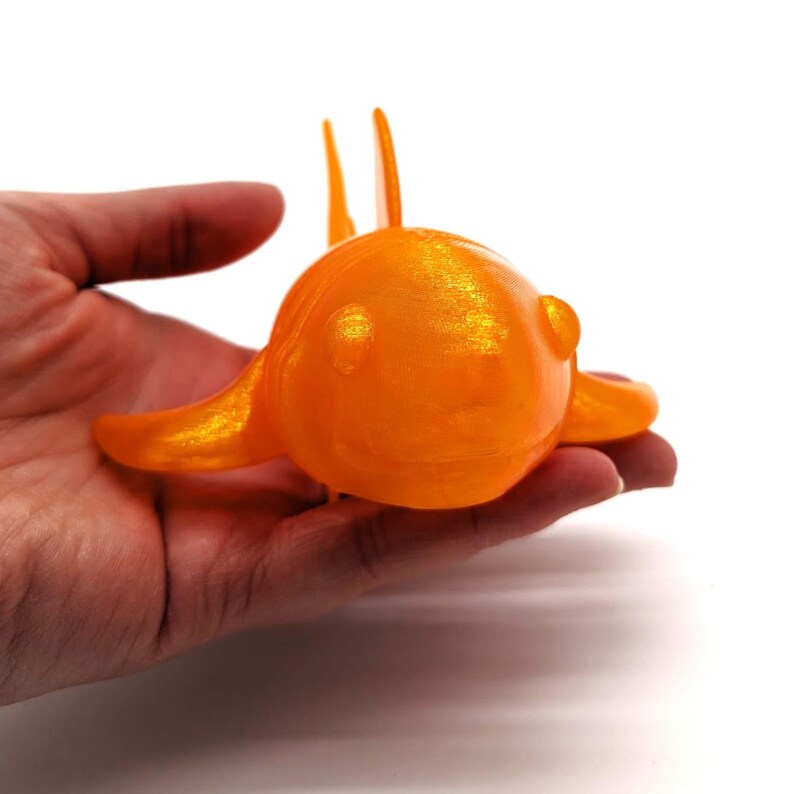 May include: An orange, fish-shaped decorative object with a shiny, metallic finish. The fish has a rounded body, fins, and two vertical protrusions. The object is held in a hand against a white background.