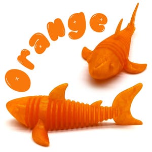 May include: Two orange shark-shaped toys with fins and segmented bodies are displayed on a white background. The word "ORANGE" is written above the sharks in orange bubble letters. One shark is positioned horizontally, and the other is vertical.