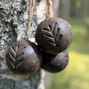 Può includere: Tre oggetti sferici marrone scuro con un disegno a foglia intagliato sulla loro superficie, appesi a una corda di fibra naturale. Gli oggetti sono raggruppati, con uno sfondo verde sfocato.