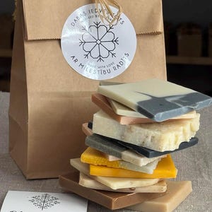 May include: A stack of handmade soap bars in various colours and patterns, including brown, yellow, white, and grey. The soaps are arranged in a tower and are packaged in a brown paper bag with a white snowflake sticker that reads "Dabas Iedvesmoti" and "Ar Milestibu Radits".