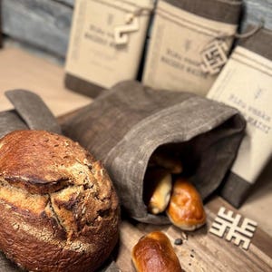 May include: A loaf of crusty brown bread and several golden-brown rolls sit on a wooden cutting board next to a linen bread bag. Several linen bread storage bags are in the background.