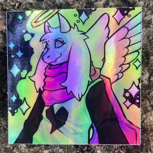 May include: A square holographic sticker featuring a stylized anthropomorphic goat character with a halo, wings, and a scarf. The character has a dark heart emblem on the chest, surrounded by colorful star shapes and a vibrant, shifting rainbow light effect.