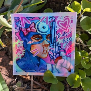 May include: Square sticker with a pop art style illustration of a person in a blue mask and bow, holding a revolver. The sticker has a holographic finish with pink and blue accents, and the words "Bulls Eye".
