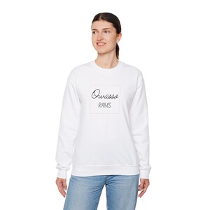 May include: A white crew neck sweatshirt featuring the text "Owasso RAMS" in black cursive script within a square. The sweatshirt is worn by a person, and is paired with blue jeans.