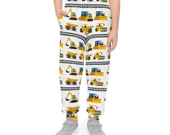 May include: White sweatpants featuring a repeating pattern of yellow construction vehicles, including excavators, dump trucks, and bulldozers. Black and white striped accents separate the rows of vehicles. The pants are worn by a person.