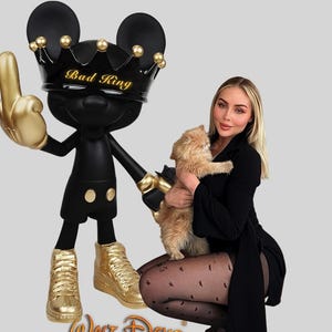 Può includere: Statuetta nera ispirata a Mickey Mouse con una corona d'oro che recita "Bad King". La statuetta alza il dito medio d'oro. Indossa scarpe da ginnastica dorate. La statuetta è accanto a una persona che tiene un gatto.