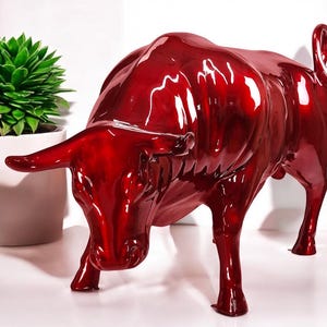 Op de afbeelding: Een glanzend, rood stierenbeeld met een gepolijste afwerking. De stier staat in een aanvallende houding, met zijn kop naar beneden en hoorns naar voren. Het beeld is een decoratief item, waarschijnlijk voor een huis of kantoor.