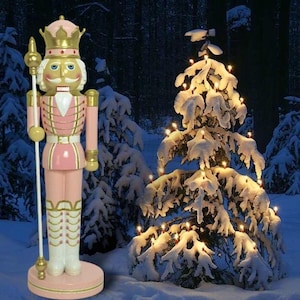 Puede incluir: Un cascanueces de madera rosa y dorado con una corona y una lanza. El cascanueces está de pie frente a un pino nevado con luces blancas.