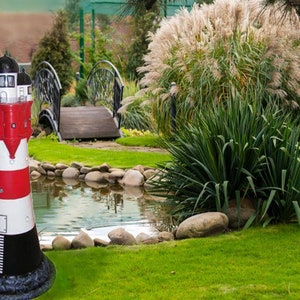 Op de afbeelding: Een rood-wit gestreept vuurtorenbeeld met een zwarte basis, dat in een tuin staat bij een vijver en een kleine brug.