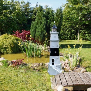 Könnte beinhalten: Ein dekorativer Leuchtturm in Schwarz und Weiß steht in einem Garten. Der Leuchtturm hat ein Licht an der Spitze und eine kleine Tür. Der Garten verfügt über einen Teich, eine Brücke und üppiges Grün.