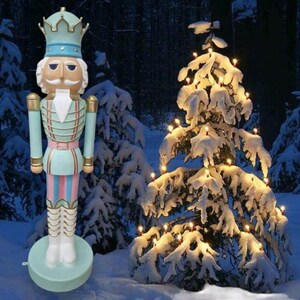 Könnte beinhalten: Eine große, blaue und rosafarbene Nussknackerfigur steht vor einem schneebedeckten Weihnachtsbaum mit weißen Lichtern. Der Nussknacker trägt eine blaue und goldene Uniform mit einem rosa-weiß gestreiften Band. Der Nussknacker hat einen weißen Bart und eine Krone auf dem Kopf.