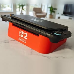 May include: A black handheld gaming console resting horizontally on a bright orange charging dock. The dock features the white Nintendo Switch logo and the number 2 printed on its front side. The console has black controllers attached.