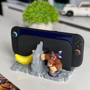 Könnte beinhalten: Eine schwarze Nintendo Switch-Konsole ruht auf einem grau-gelben Donkey Kong-Ständer. Der Ständer zeigt eine geformte Donkey Kong-Figur mit einem Fass und einer Banane. Die Konsole hat blaue und schwarze Controller.
