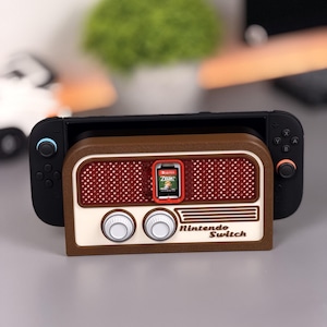 Pode incluir: Uma base Nintendo Switch castanha e creme com um design de rádio retro. A base contém uma consola Nintendo Switch e um cartucho de jogo. O texto "Nintendo Switch" é visível na frente.