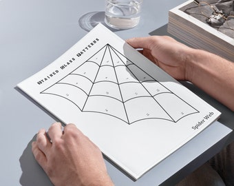 Puede incluir: Una hoja de papel blanca con un diseño de telaraña en blanco y negro. El papel tiene el título "Stained Glass Patterns" y las palabras "Spider Web" en la parte inferior. El papel está siendo sostenido por las manos de una persona.