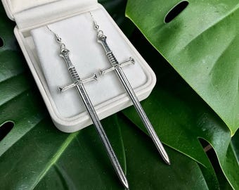 May include: A pair of silver-toned sword earrings featuring detailed hilts, crossguards, and long blades. The earrings are suspended from standard ear wires and are displayed inside a white jewelry box with a soft, light-colored interior lining.