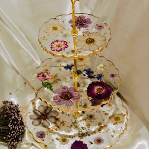 K&ouml;nnte beinhalten: Ein dreist&ouml;ckiger Kuchenst&auml;nder mit einem goldenen Metallrahmen und gewellten Kanten. Jede Etage ist mit gepressten Blumen und goldenen Blattverzierungen verziert.