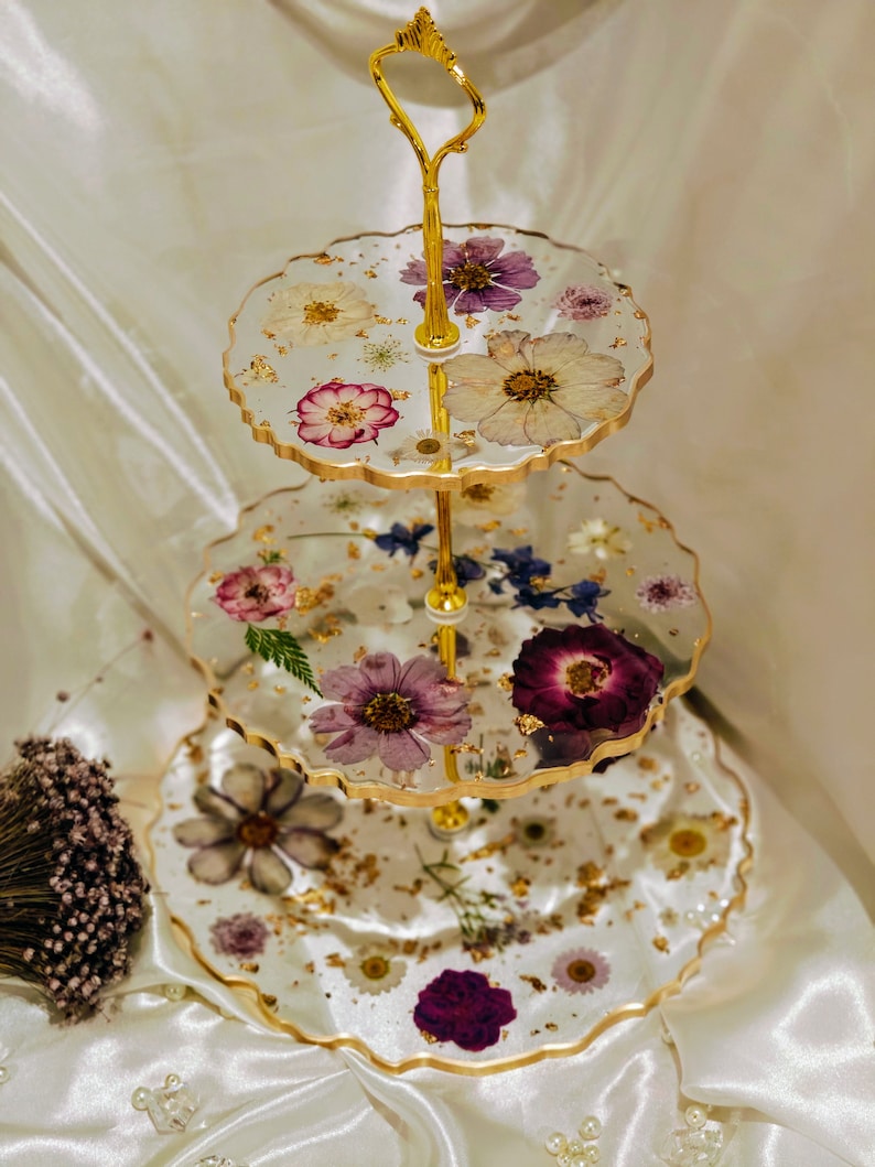 K&ouml;nnte beinhalten: Ein dreist&ouml;ckiger Kuchenst&auml;nder mit einem goldenen Metallrahmen und gewellten Kanten. Jede Etage ist mit gepressten Blumen und goldenen Blattverzierungen verziert.