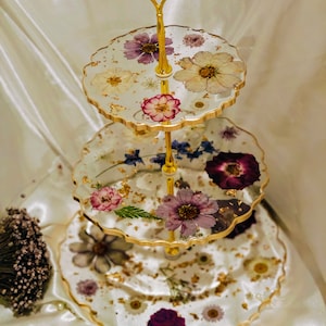 K&ouml;nnte beinhalten: Ein dreist&ouml;ckiger Kuchenst&auml;nder mit einem goldenen Metallrahmen und durchsichtigen Harztabletts. Jedes Tablett ist mit gepressten Blumen und Blattgold verziert. Der St&auml;nder ist perfekt zum Pr&auml;sentieren von Desserts oder anderen Leckereien.