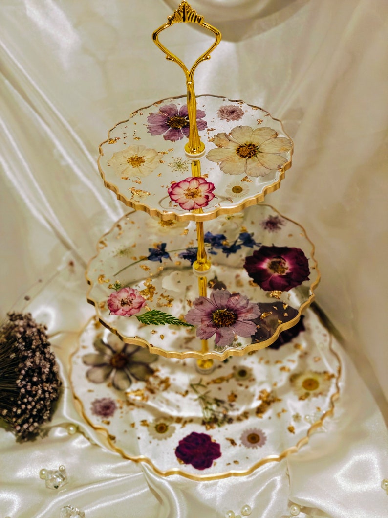 K&ouml;nnte beinhalten: Ein dreist&ouml;ckiger Kuchenst&auml;nder mit einem goldenen Metallrahmen und durchsichtigen Harztabletts. Jedes Tablett ist mit gepressten Blumen und Blattgold verziert. Der St&auml;nder ist perfekt zum Pr&auml;sentieren von Desserts oder anderen Leckereien.
