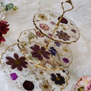 K&ouml;nnte beinhalten: Ein dreist&ouml;ckiger Kuchenst&auml;nder mit einem goldenen Metallrahmen und drei runden Glasplatten, die mit gepressten Blumen und Blattgold verziert sind. Die Platten sind klar und zeigen die Blumen und das Blattgold durch das Glas.