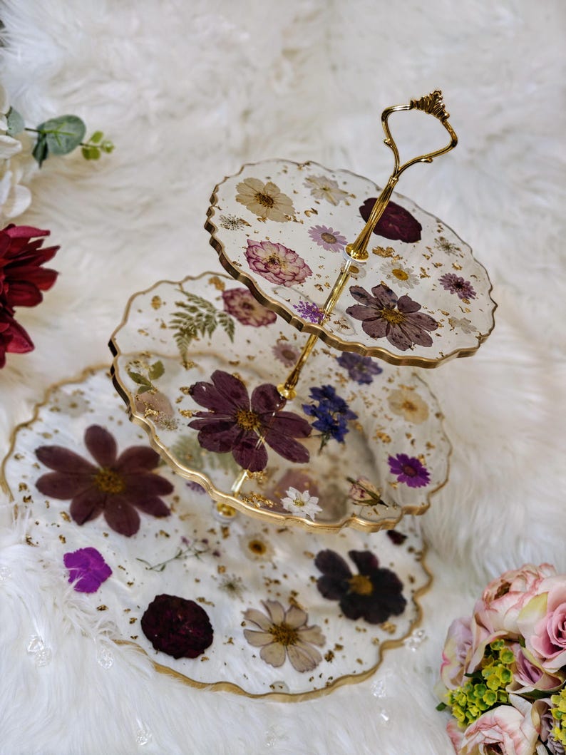 K&ouml;nnte beinhalten: Ein dreist&ouml;ckiger Kuchenst&auml;nder mit einem goldenen Metallrahmen und drei runden Glasplatten, die mit gepressten Blumen und Blattgold verziert sind. Die Platten sind klar und zeigen die Blumen und das Blattgold durch das Glas.
