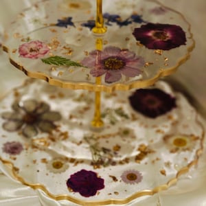K&ouml;nnte beinhalten: Ein zweist&ouml;ckiger Kuchenst&auml;nder mit einem goldenen Metallrahmen und klaren Glasplatten. Die Platten sind mit gepressten Blumen und Blattgold verziert.