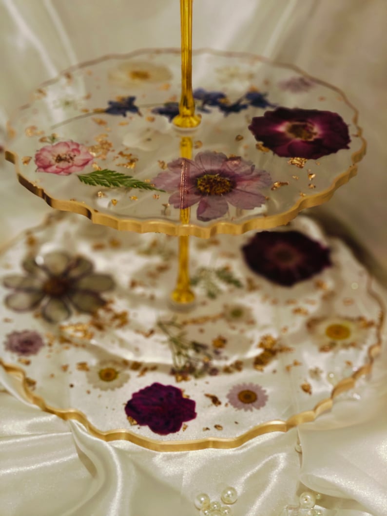 K&ouml;nnte beinhalten: Ein zweist&ouml;ckiger Kuchenst&auml;nder mit einem goldenen Metallrahmen und klaren Glasplatten. Die Platten sind mit gepressten Blumen und Blattgold verziert.