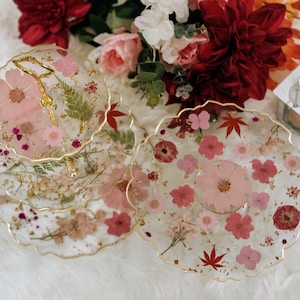 May include: A three-tiered cake stand with a gold metal frame and delicate floral accents. Each tier is made of clear resin with pressed flowers and gold leaf accents.