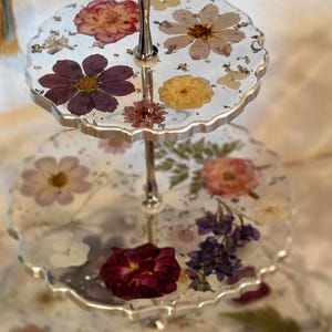 K&ouml;nnte beinhalten: Ein dreist&ouml;ckiger Silberst&auml;nder mit einem gewellten Rand. Jede Etage ist mit gepressten Blumen in Rosa-, Lila- und Gelbt&ouml;nen gef&uuml;llt.