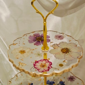 K&ouml;nnte beinhalten: Ein zweist&ouml;ckiger Kuchenst&auml;nder mit einem goldenen Metallrahmen und Griff. Die obere Etage hat einen wei&szlig;en Hintergrund mit goldenen Blattverzierungen und gepressten Blumen in Violett-, Wei&szlig;- und Rosat&ouml;nen. Die untere Etage hat ein &auml;hnliches Design mit blauen und violetten Blumen.